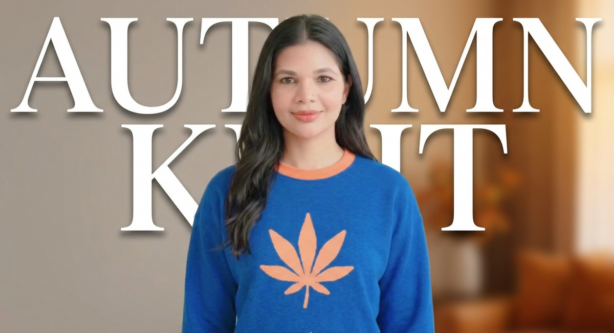 A woman wearing a blue crewneck sweater with a peach leaf graphic, standing in front of a blurred indoor background with large white text reading 'AUTUMN KNOT'.